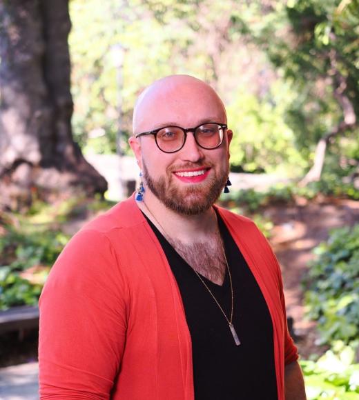 a smiling individual with a beard and glasses, wearing a black top and pink sweater